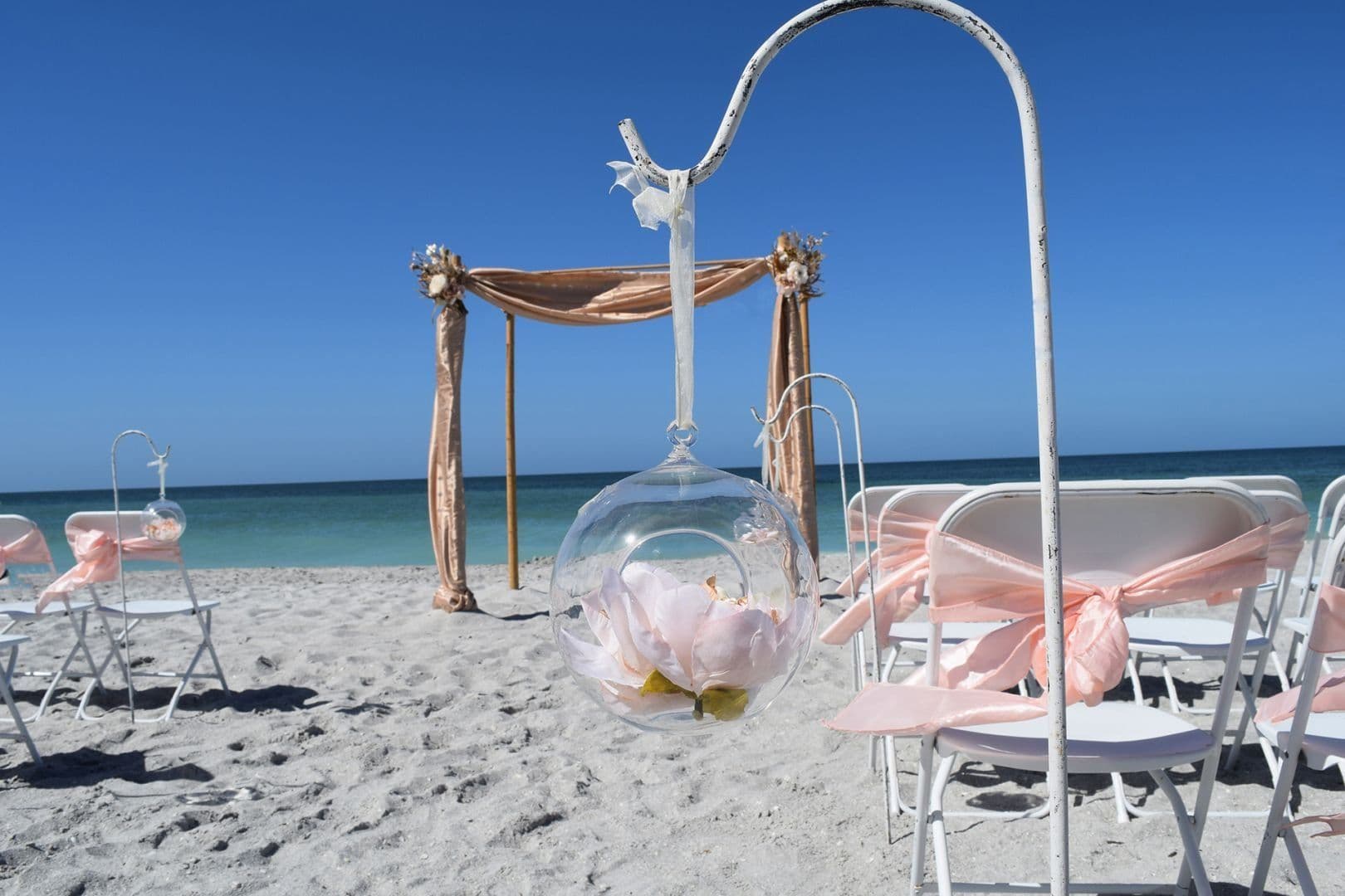 Beach wedding setup: chairs with pink bows, floral decor, draped arch, clear blue sky.